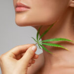 A women gently holds a green cannabis leaf near their neck, subtly suggesting the soothing benefits of CBD in skincare. The image focuses on the lower half of the face and hand, set against a soft, neutral background that highlights this serene moment.
