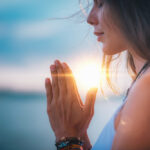 A woman stands by the water, her hands pressed together in a prayer-like position as part of her self-care practices. The sun sets behind her, casting a warm glow. She appears peaceful, with her hair gently blowing in the breeze against a serene, blue sky backdrop.
