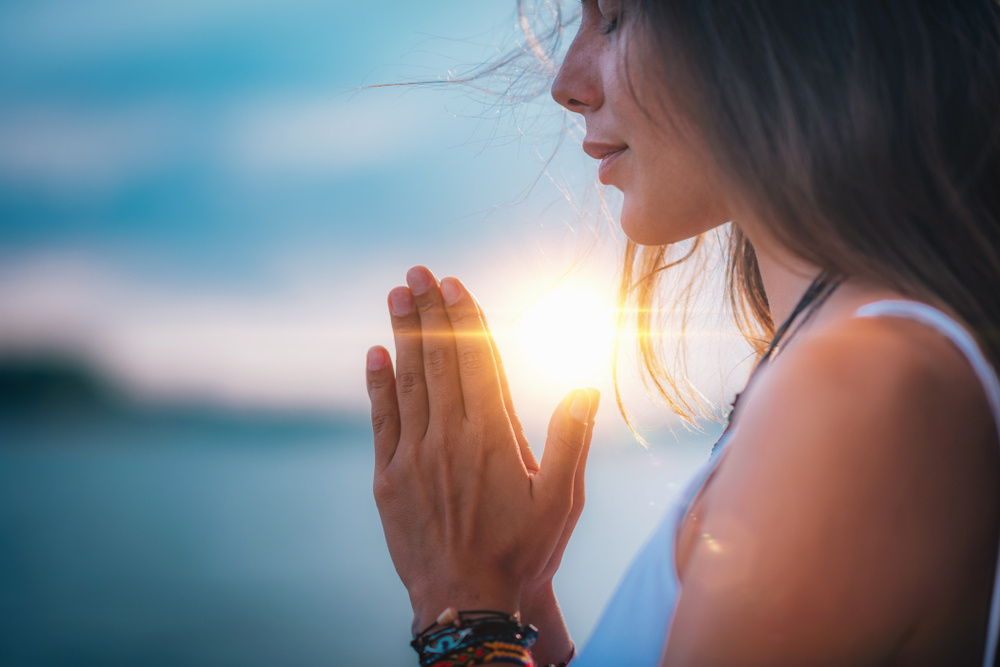 A woman stands by the water, her hands pressed together in a prayer-like position as part of her self-care practices. The sun sets behind her, casting a warm glow. She appears peaceful, with her hair gently blowing in the breeze against a serene, blue sky backdrop.