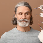A middle-aged man with gray hair and a beard looks skeptically at several syringes, reminiscent of a botox guide, pointed towards his face. He is wearing a gray shirt, and gloved hands hold the syringes against a brown background.
