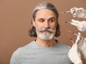 A middle-aged man with gray hair and a beard looks skeptically at several syringes, reminiscent of a botox guide, pointed towards his face. He is wearing a gray shirt, and gloved hands hold the syringes against a brown background.