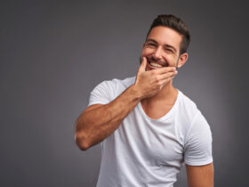A man with short dark hair and a beard is smiling warmly, touching his chin with his left hand as if contemplating skin care. He is wearing a white T-shirt and stands against a plain gray background.