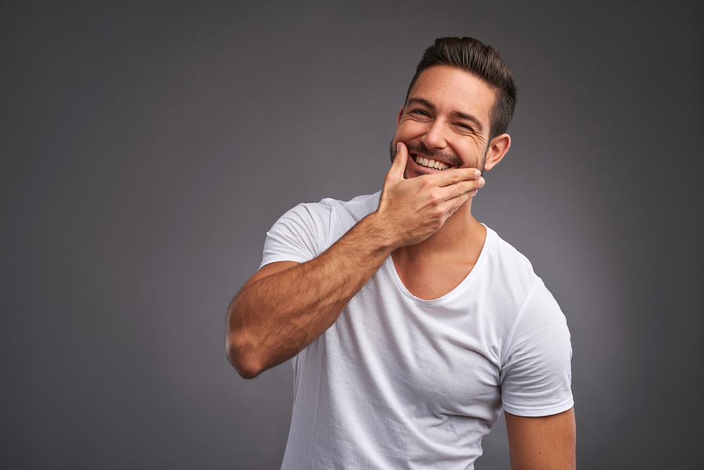 A man with short dark hair and a beard is smiling warmly, touching his chin with his left hand as if contemplating skin care. He is wearing a white T-shirt and stands against a plain gray background.