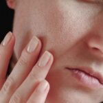 Close-up of a person's face, highlighting the skin pores. The fingers gently touch the cheek, emphasizing the texture and natural complexion beautifully illuminated by the light.