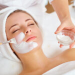A person is lying down with a towel wrapped around their head, indulging in a luxurious skincare treatment. A skincare professional carefully applies a white face mask using a brush, expertly holding a small bowl of the soothing mask mixture.