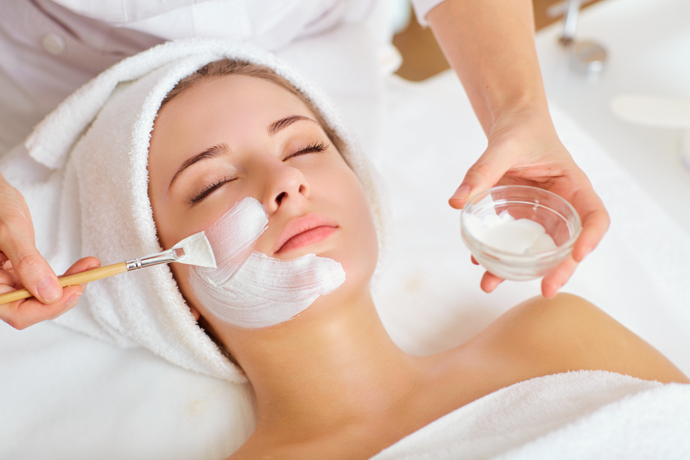 A person is lying down with a towel wrapped around their head, indulging in a luxurious skincare treatment. A skincare professional carefully applies a white face mask using a brush, expertly holding a small bowl of the soothing mask mixture.