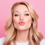 A person with long blonde hair poses against a pink background, making a playful kissing face with smooth lips. They are wearing a light-colored top.