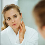 A woman in a white bathrobe looks at herself in the mirror, gently touching her face with one hand. The reflection reveals her focused expression, noticing faint white spots as she inspects her skin. In the background, everything is softly blurred.