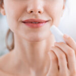 A close-up of a person enhancing the look of your lips by applying lip balm. The tube is held near their mouth, highlighting the smoothness and care given to the lips. The background remains softly blurred, drawing focus to this intimate moment of self-care.