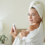 A person wearing a white bathrobe and a towel on their head smiles, embracing self-care, while holding a small bottle. They stand in a softly lit bedroom with a lamp and a potted plant in the background.