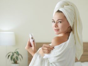 A person wearing a white bathrobe and a towel on their head smiles, embracing self-care, while holding a small bottle. They stand in a softly lit bedroom with a lamp and a potted plant in the background.