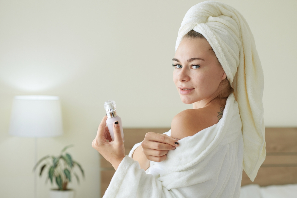 A person wearing a white bathrobe and a towel on their head smiles, embracing self-care, while holding a small bottle. They stand in a softly lit bedroom with a lamp and a potted plant in the background.