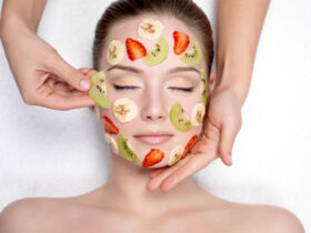 A person with fruit slices, including kiwi, strawberry, and banana, placed on their face as part of a facial treatment for glowing skin. Hands are gently adjusting the slices while the person lies with their eyes closed, showing a relaxed expression.