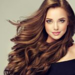 A woman with long, flowing brown hair wears an off-the-shoulder black top and smiles softly. Her long locks frame her bright eyes and radiant complexion, set against a neutral background.