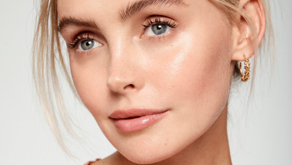 Close-up of a person with light skin, blue eyes, and blonde hair. They wear a gold hoop earring, showcasing their glowing skin with a natural makeup look. The softly blurred background adds an exquisite touch to their radiant appearance.