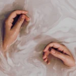 Two hands partially submerged in a milky, swirling liquid, practicing skin flooding. The creamy texture of the liquid creates an artistic, calming effect around the hands.