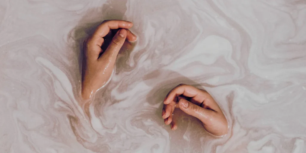 Two hands partially submerged in a milky, swirling liquid, practicing skin flooding. The creamy texture of the liquid creates an artistic, calming effect around the hands.