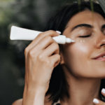 A woman is applying sunscreen to her face from a tube while smiling with her eyes closed. She is surrounded by greenery, creating a natural and serene setting.