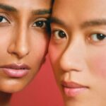Close-up of two individuals with pink blush and natural makeup, embodying beauty against a red backdrop. One has long brown hair, the other short, light-colored hair. They wear gold earrings and their faces are slightly touching, capturing the essence that beauty brands strive to achieve.