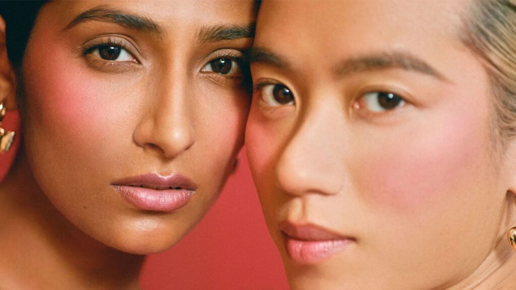Close-up of two individuals with pink blush and natural makeup, embodying beauty against a red backdrop. One has long brown hair, the other short, light-colored hair. They wear gold earrings and their faces are slightly touching, capturing the essence that beauty brands strive to achieve.