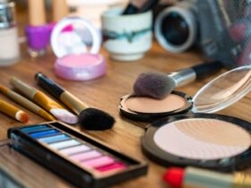 The wooden table is adorned with an array of makeup essentials like brushes, eyeshadow palettes, blush, and lipstick. A compact powder sits elegantly among the collection, while a camera lens peeks from the background. The vibrant scene is beautifully lit, capturing the artistry of makeup.