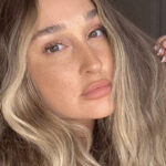 A person with long, wavy hair and natural highlights poses indoors, exuding a sense of natural beauty. They have a neutral expression and gently touch their hair with one hand. The softly lit background features a visible door.