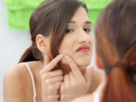 A woman with brunette hair examines her face in the mirror, her fingers gently checking for any signs of acne. She appears focused, with a slightly serious expression. Green and white towels hang in the background, adding a soft touch to the scene.