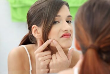 A woman with brunette hair examines her face in the mirror, her fingers gently checking for any signs of acne. She appears focused, with a slightly serious expression. Green and white towels hang in the background, adding a soft touch to the scene.