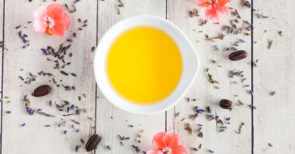 A white bowl filled with yellow liquid sits on a wooden surface, surrounded by scattered dried lavender, pink flowers, and coffee beans. This serene scene exudes a rustic charm, embodying the soothing essence of natural hair care rituals.
