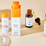 A collection of skincare products is displayed. From left to right: a bowl with cotton pads, two facial serums in orange containers, a dropper bottle, and a cream with a pump dispenser. A glass surface with water droplets is in the foreground.