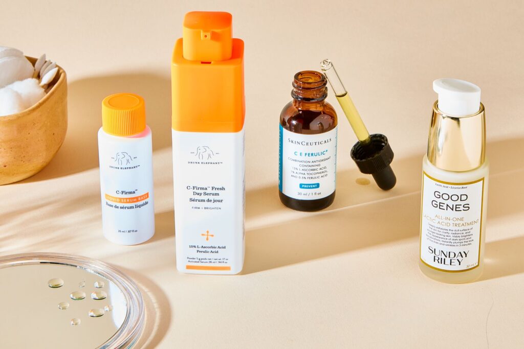 A collection of skincare products is displayed. From left to right: a bowl with cotton pads, two facial serums in orange containers, a dropper bottle, and a cream with a pump dispenser. A glass surface with water droplets is in the foreground.