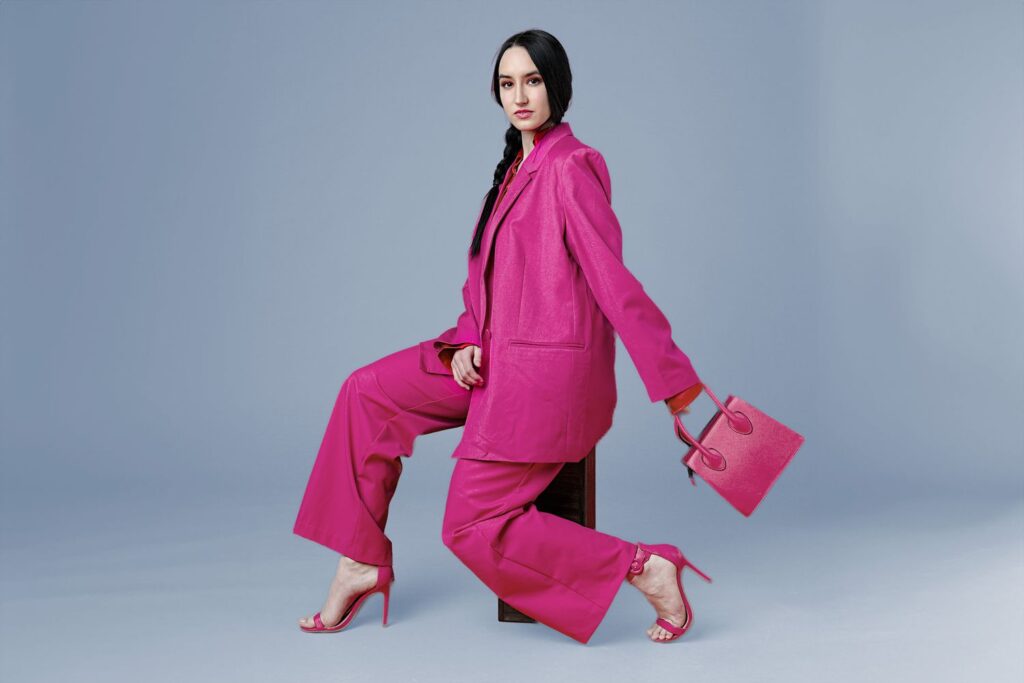 A person wearing a fashion-forward bright pink suit and heels sits on a stool against a light gray background. They hold a matching pink handbag, with hair in a long braid, embodying the essence of this season's fashion forecast.