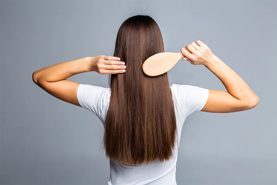 A person with long, straight brown hair is engaged in hair care, brushing their locks with a wooden brush. They are wearing a white shirt and facing away from the camera against a plain gray background.