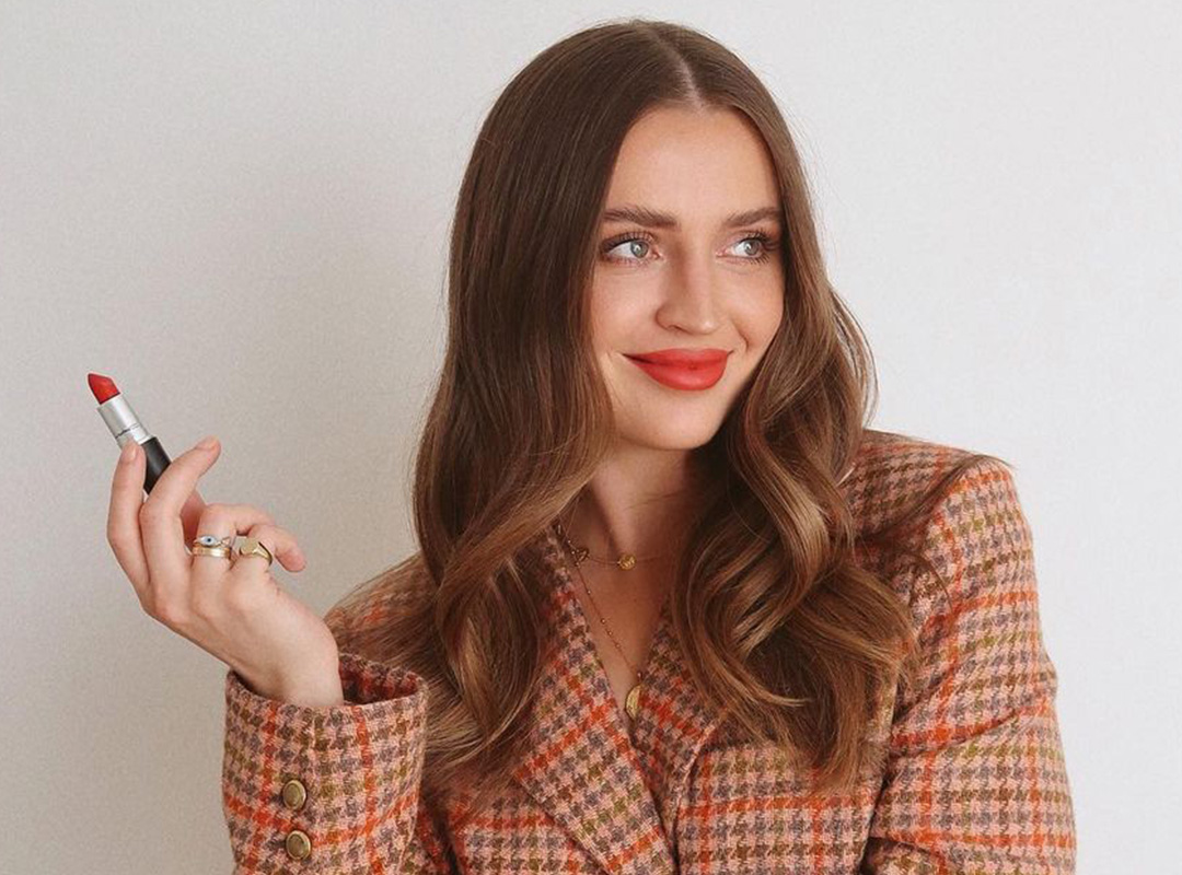 A person with long brown hair is smiling and holding a red lipstick, showcasing their makeup transformation. They are wearing a checkered blazer in shades of pink and orange against a plain background.