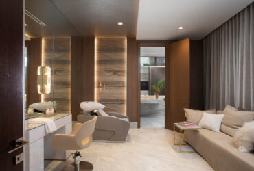A modern salon with a sleek interior design features a styling station complete with mirror and chair, evoking a home spa vibe. A comfortable beige sofa with cushions and a small side table with books enrich the experience. The well-lit room highlights elegant wood accents, creating an inviting atmosphere.