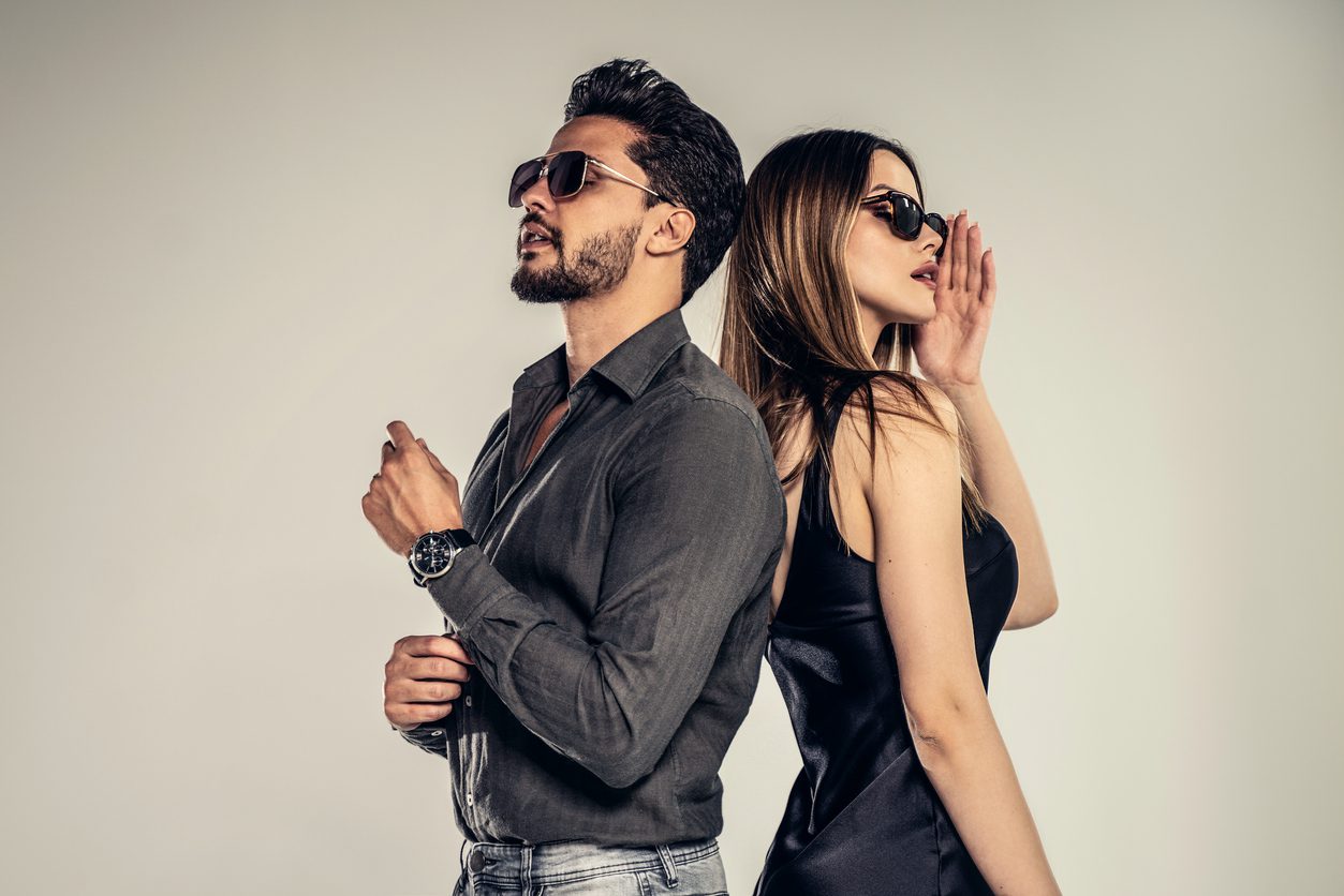 A man and a woman stand back-to-back, both wearing sunglasses in a stylish fashion. The man has a beard and wears a gray shirt with rolled-up sleeves, while the woman flaunts her long hair in a sleek black dress. Together, they exude an air of confident elegance.