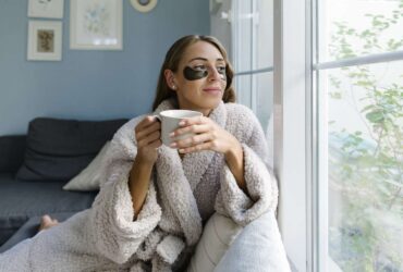 A woman in a robe enjoys coffee while looking out the window, her dark circles suggesting a need for more sleep.