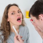 A man examines a woman's mouth with a toothbrush, focusing on her swollen uvula during a dental check-up.