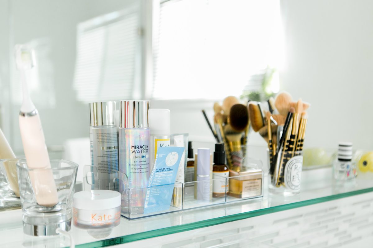 A neatly arranged collection of Makeup And Beauty Products, including skincare bottles, brushes, and an electric toothbrush, all placed on a glass shelf