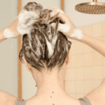 Women washing her hair in the shower with shampoo lathering on the scalp under a modern rainfall showerhead.