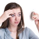 A concerned young woman examining her forehead in a small mirror, checking for blemishes and signs of irritation, highlighting the importance of knowing which ingredients to avoid putting on skin.