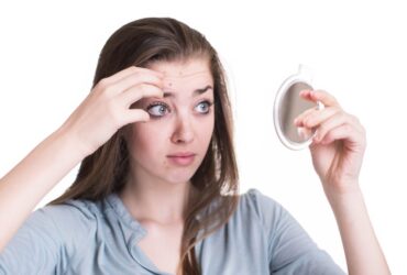 A concerned young woman examining her forehead in a small mirror, checking for blemishes and signs of irritation, highlighting the importance of knowing which ingredients to avoid putting on skin.
