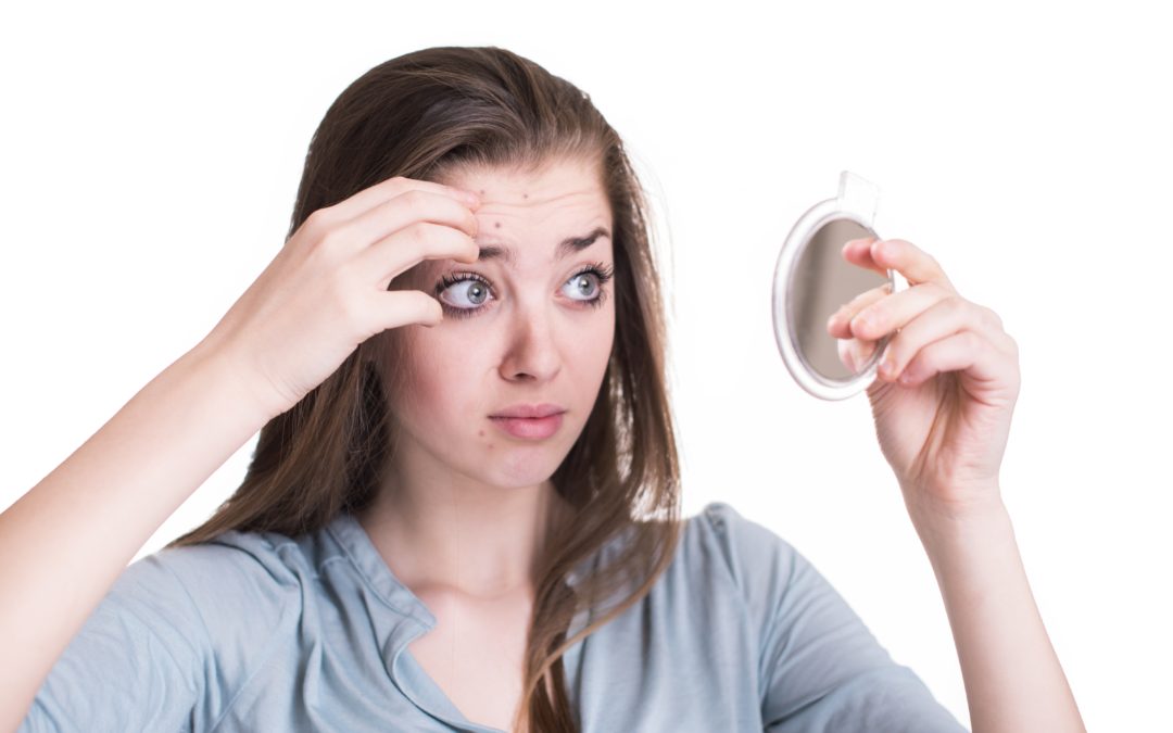 A concerned young woman examining her forehead in a small mirror, checking for blemishes and signs of irritation, highlighting the importance of knowing which ingredients to avoid putting on skin.