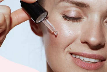 A smiling person gently applies retinol serum to their cheek with a dropper. Their eyes are closed, and their skin appears dewy and smooth, showcasing the effectiveness of their skin-care routine.